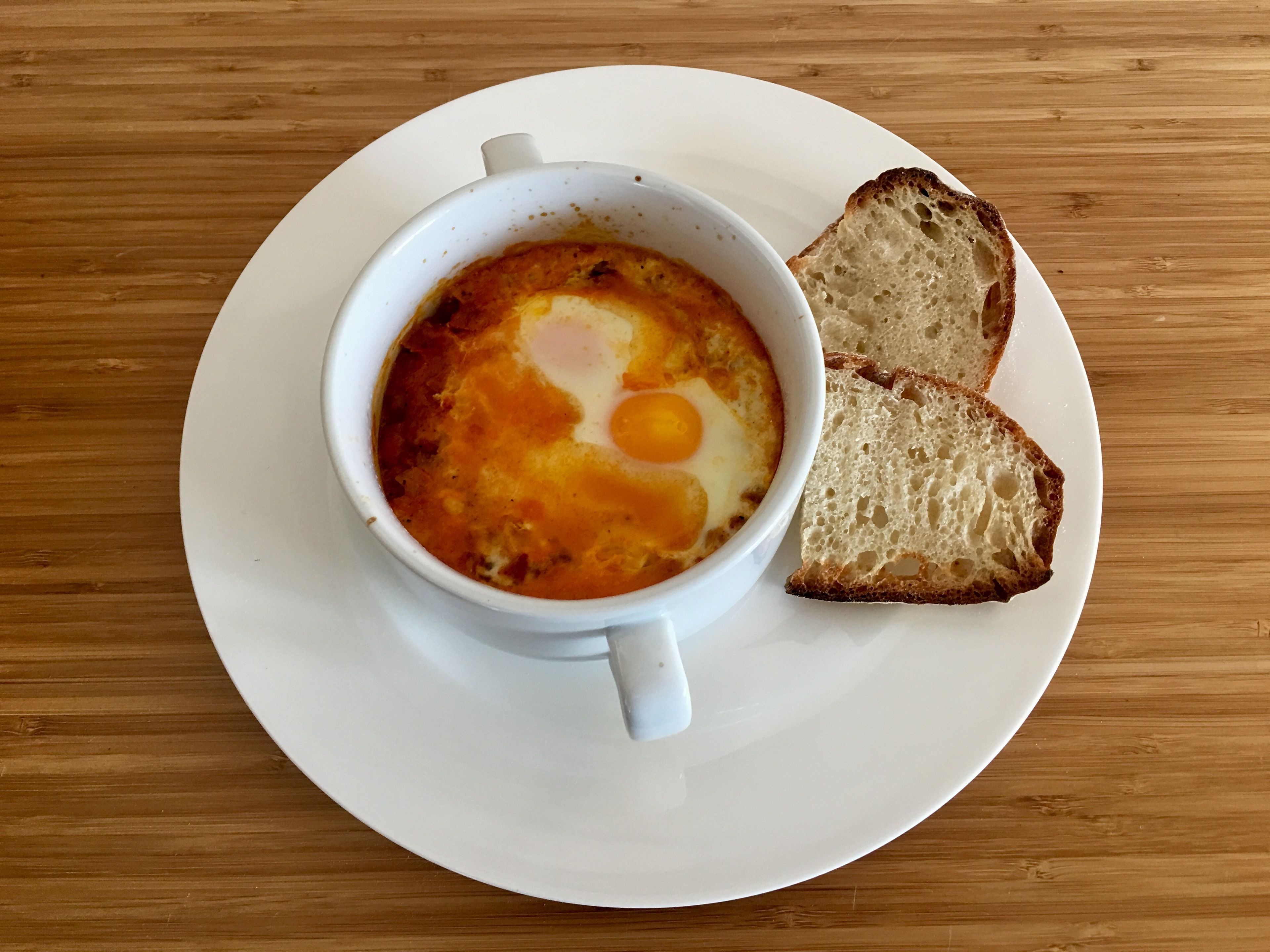 A bowl of shakshuka with eggs on a white plate, accompanied by slices of toast.