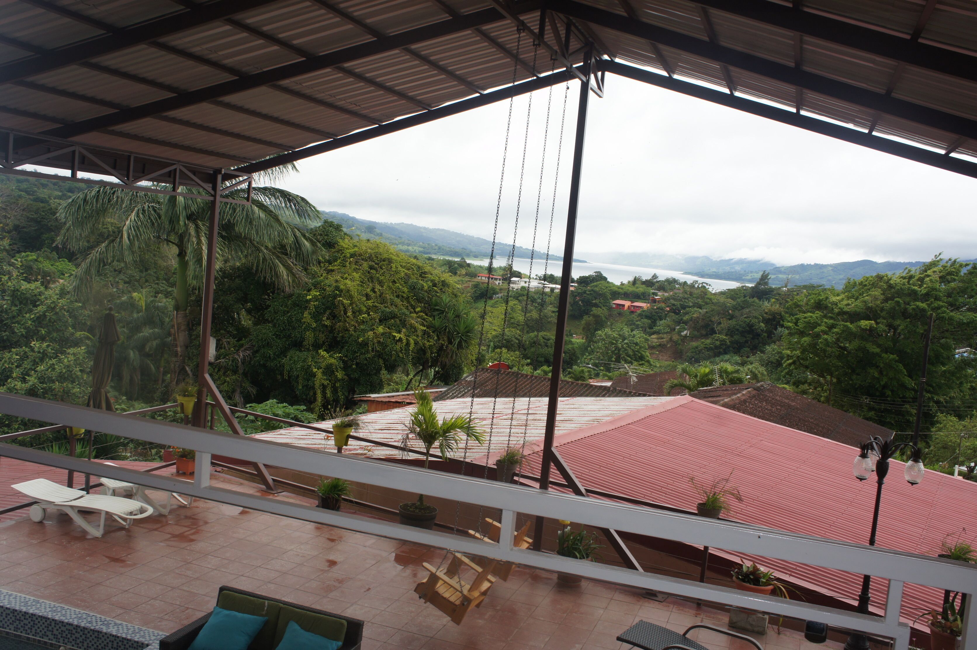 A covered outdoor area with a view of a red-roofed building, palm trees, and a mountainous landscape.