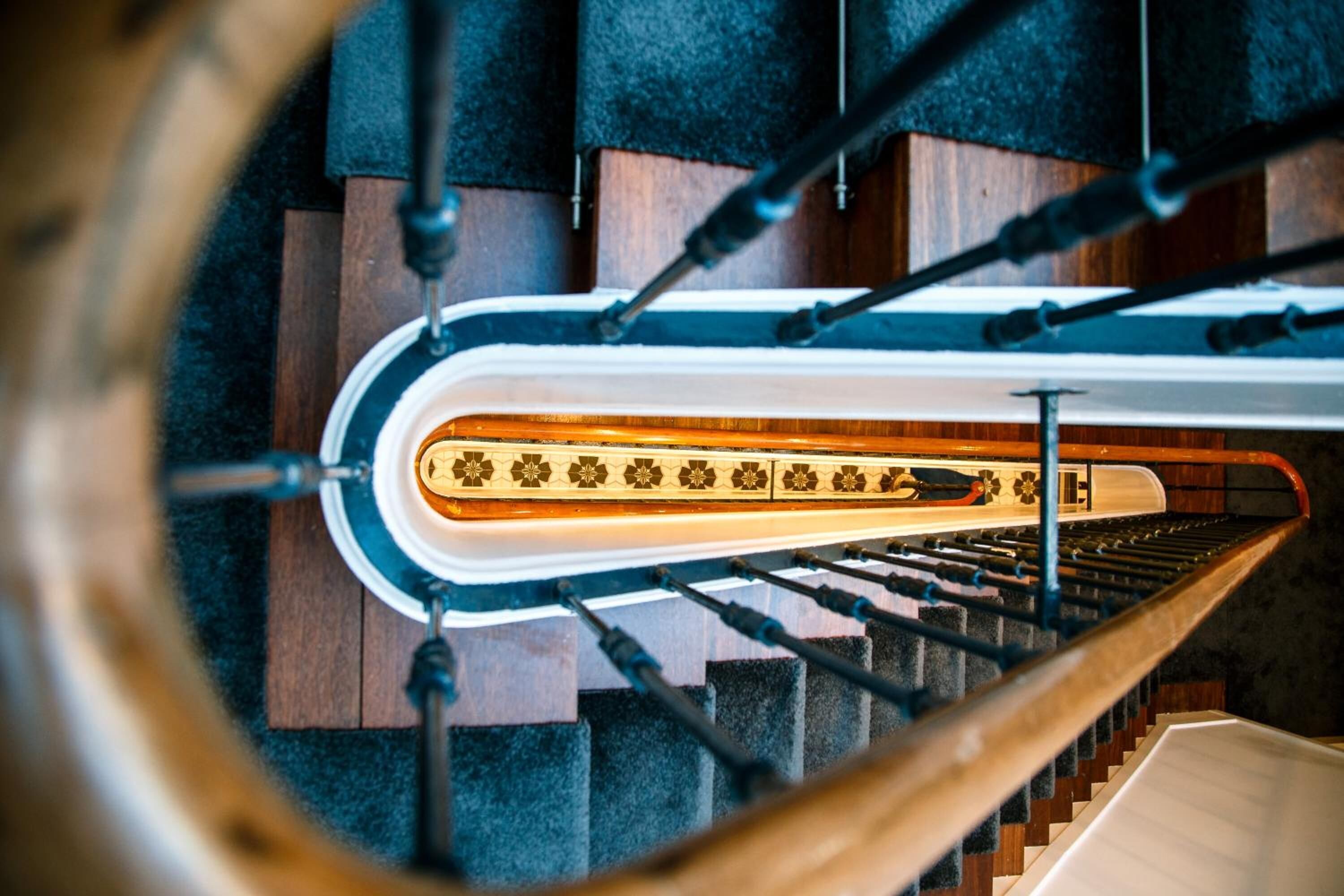A spiral staircase with a wooden handrail and black balusters, leading to a landing with a patterned floor.