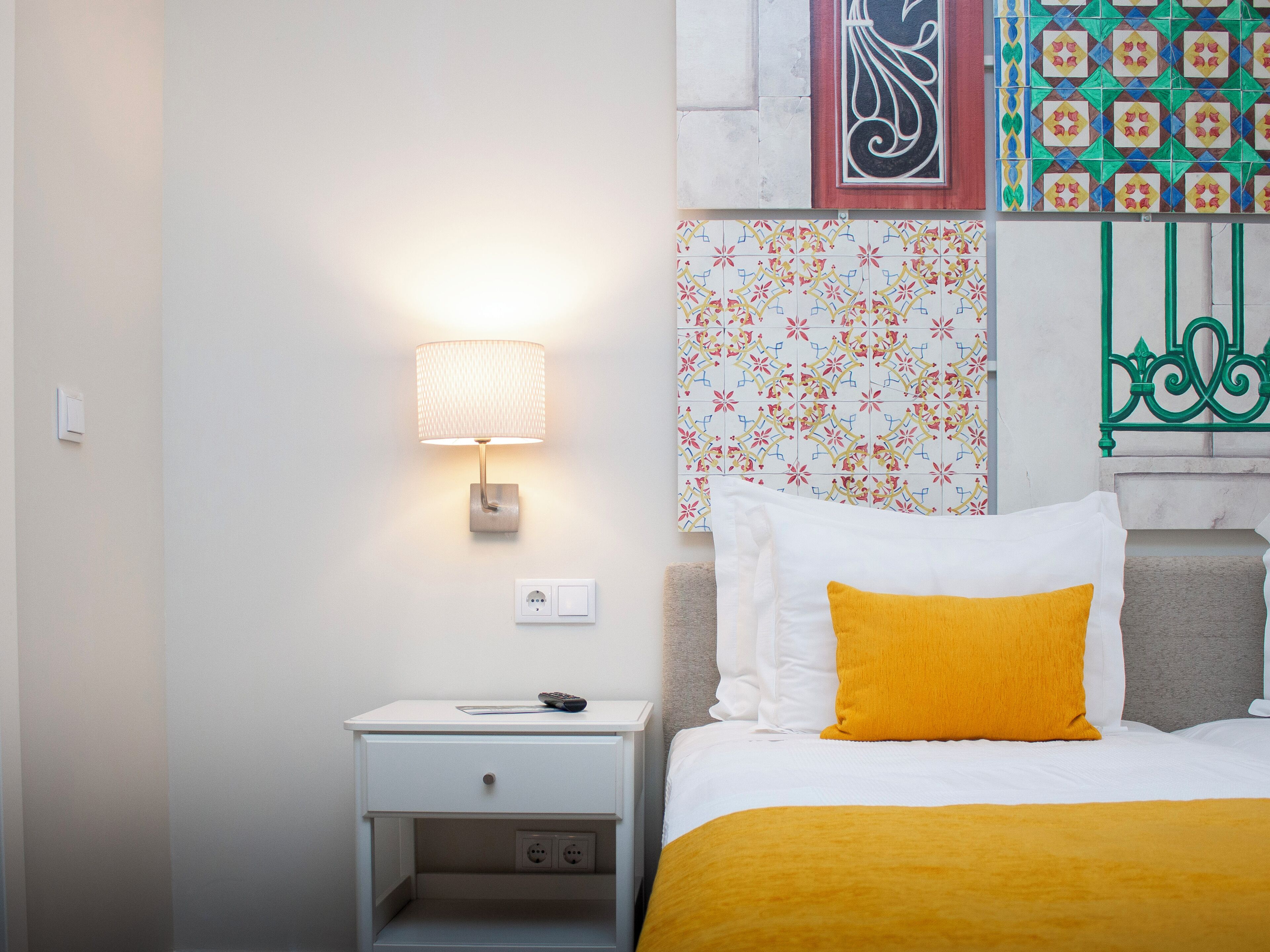 A neatly arranged hotel room with a bedside lamp, a bedside table with a remote control, a bed with white and yellow bedding, and decorative wall art.