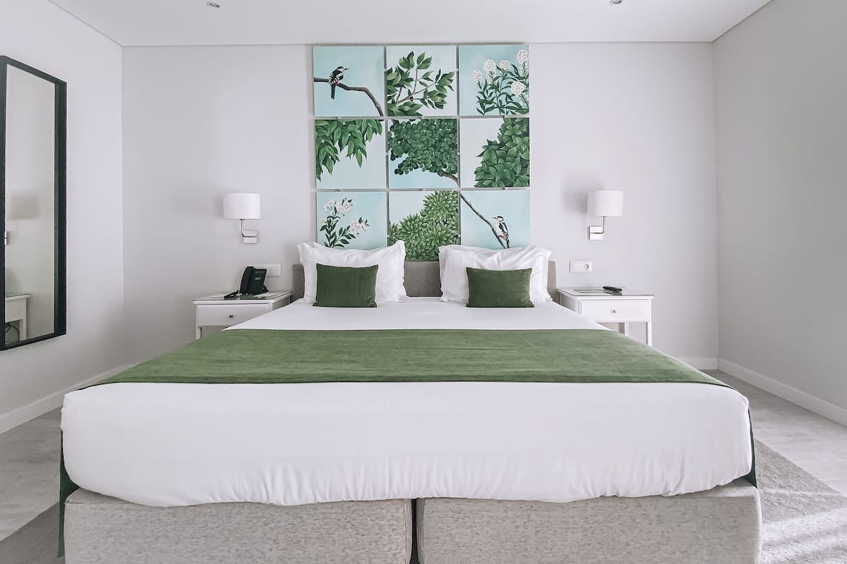 A modern hotel room with a large bed, two bedside tables, a nightstand, a mirror, and a wall with a nature-themed artwork.