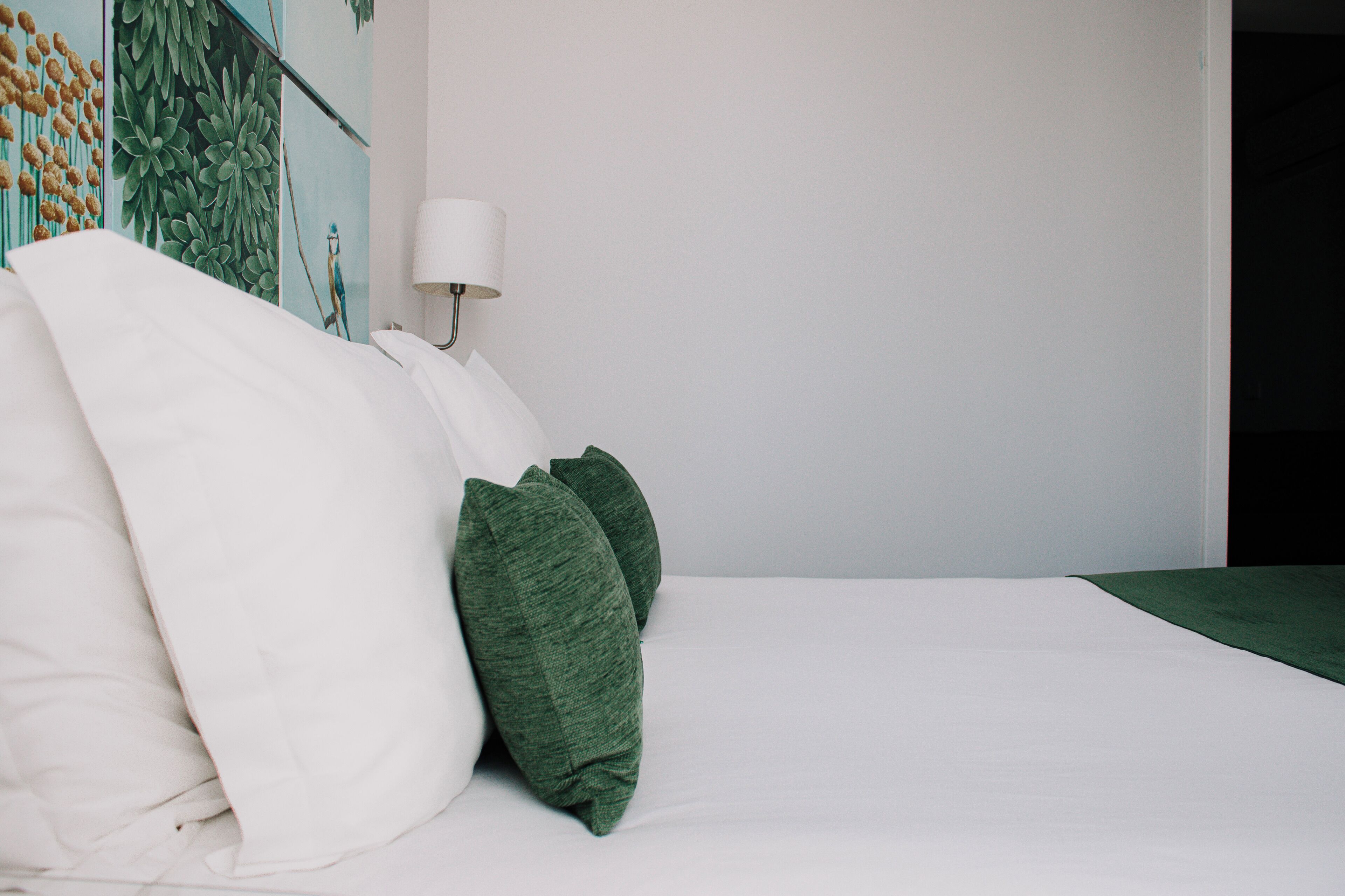 A neatly made bed with white and green pillows, a white wall, and a lamp.