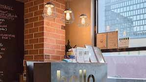 Een receptiebalie met een 'hello' bord, een bakstenen muur, hangende pendelverlichting en een raam met uitzicht op een gebouw.