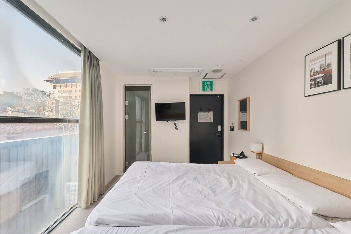 Een hotelkamer met een groot bed, een televisie, een deur met een nooduitgangsbordje en een raam met gordijnen.