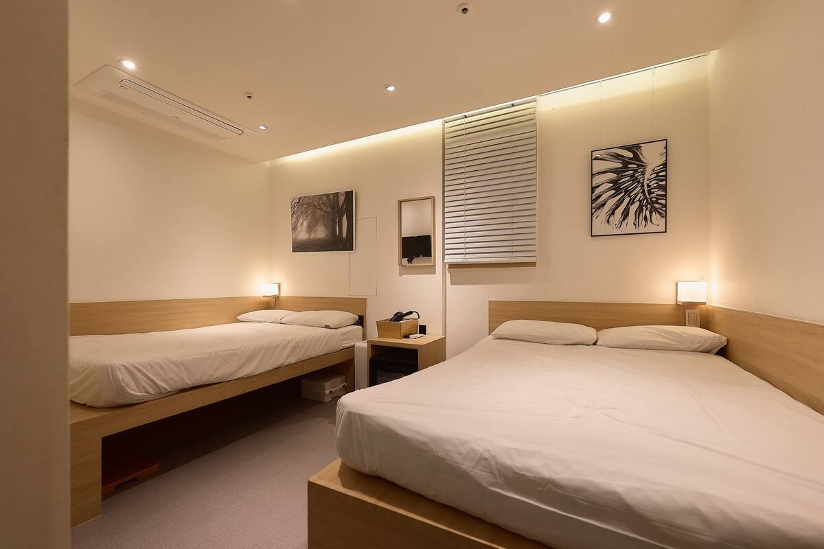 Een hotelkamer met twee bedden, een nachtkastje met lamp, een televisie aan de muur en een raam met zonwering.