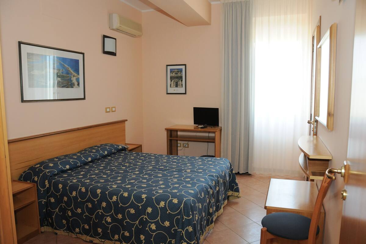 A hotel room with a bed, a desk, a chair, a television, and two framed pictures on the wall.