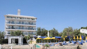 Ein mehrstöckiges Hotel mit Balkonen, ein Strandbereich mit Liegestühlen und Sonnenschirmen sowie ein kleines Pavillon.