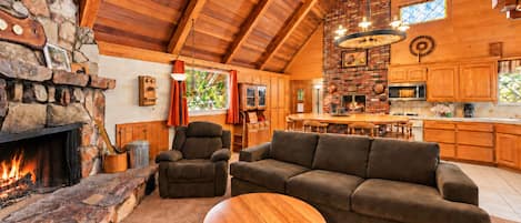 A cozy living room with a fireplace, a stone wall, and a high ceiling with wooden beams.