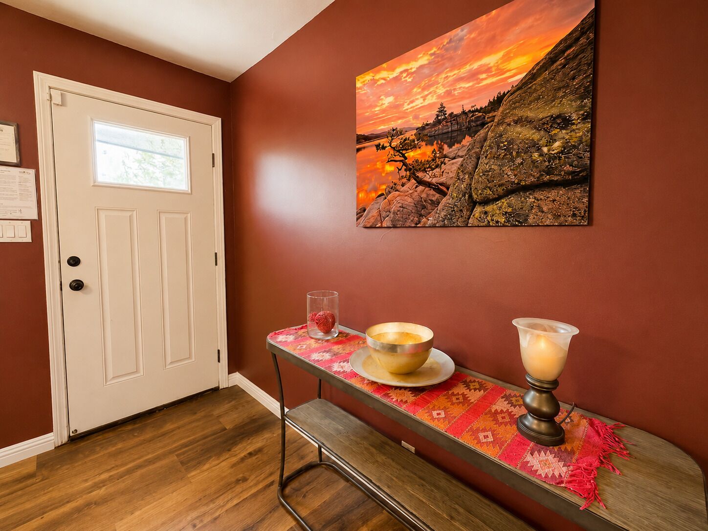 A room with a wooden floor, a white door, a painting of a sunset landscape, a table with a candle, and a glass of water.