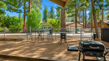 An outdoor patio area with a grill, a table, and chairs, surrounded by trees and a fence.