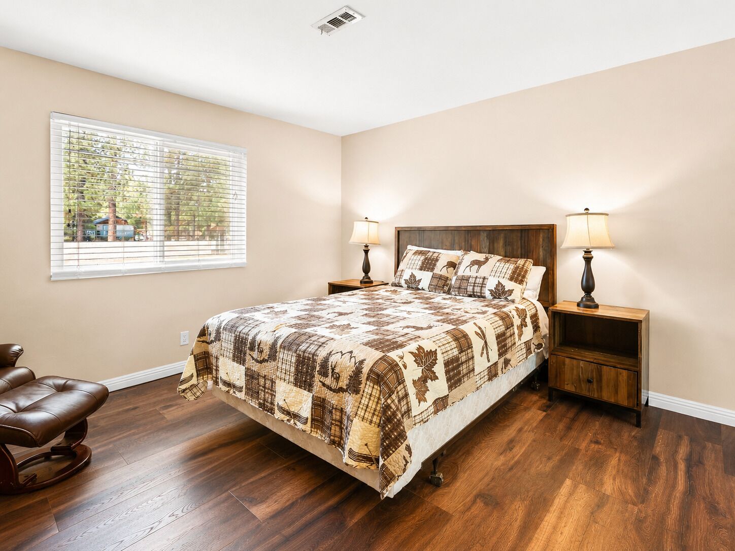 A bedroom with a large bed, two bedside tables with lamps, a window with blinds, and a brown leather chair.