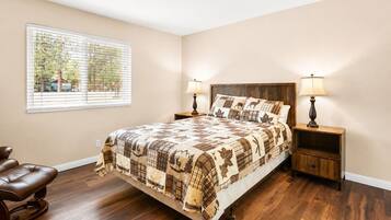 A bedroom with a large bed, two bedside tables with lamps, a window with blinds, and a brown leather chair.