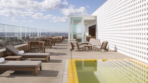 Een terras op het dak met lounge fauteuils, een zwembad en een glazen ruimte met tafels en stoelen.