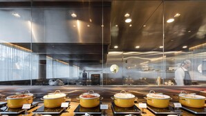 Een buffetgedeelte met meerdere gele kommen vol eten, serveerbestek en een modern interieur met glazen scheidingswanden en roestvrijstalen oppervlakken.