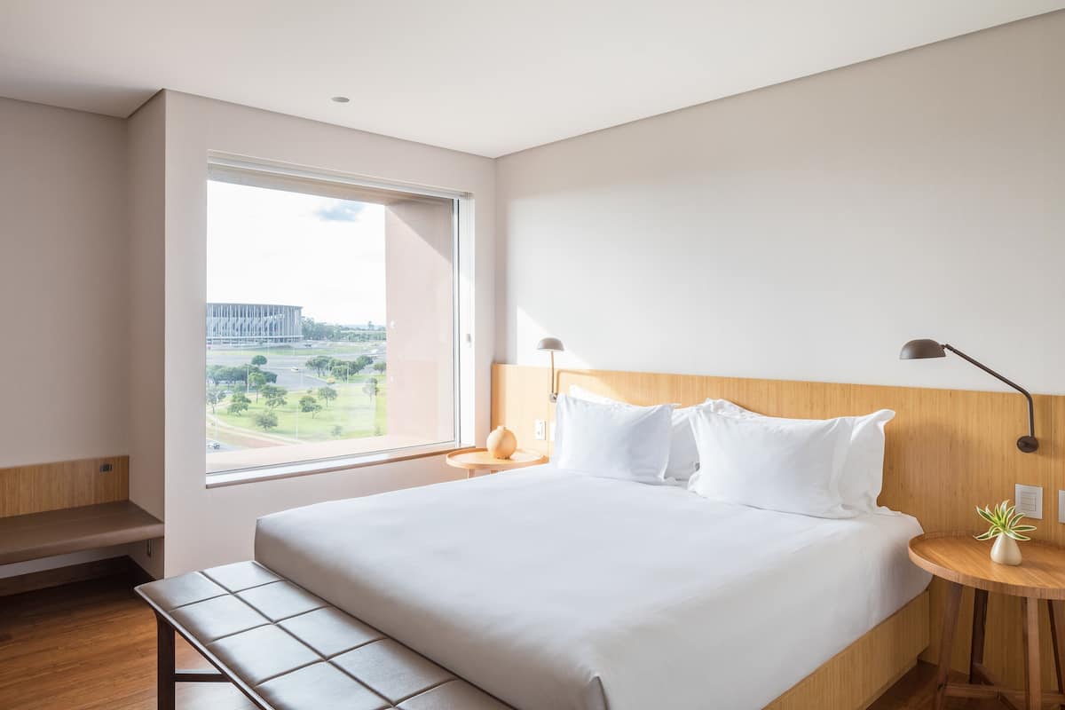 Een moderne hotelkamer met een groot bed, een bank, een ronde tafel met een lamp, en uitzicht op een gebouw en groen buiten.