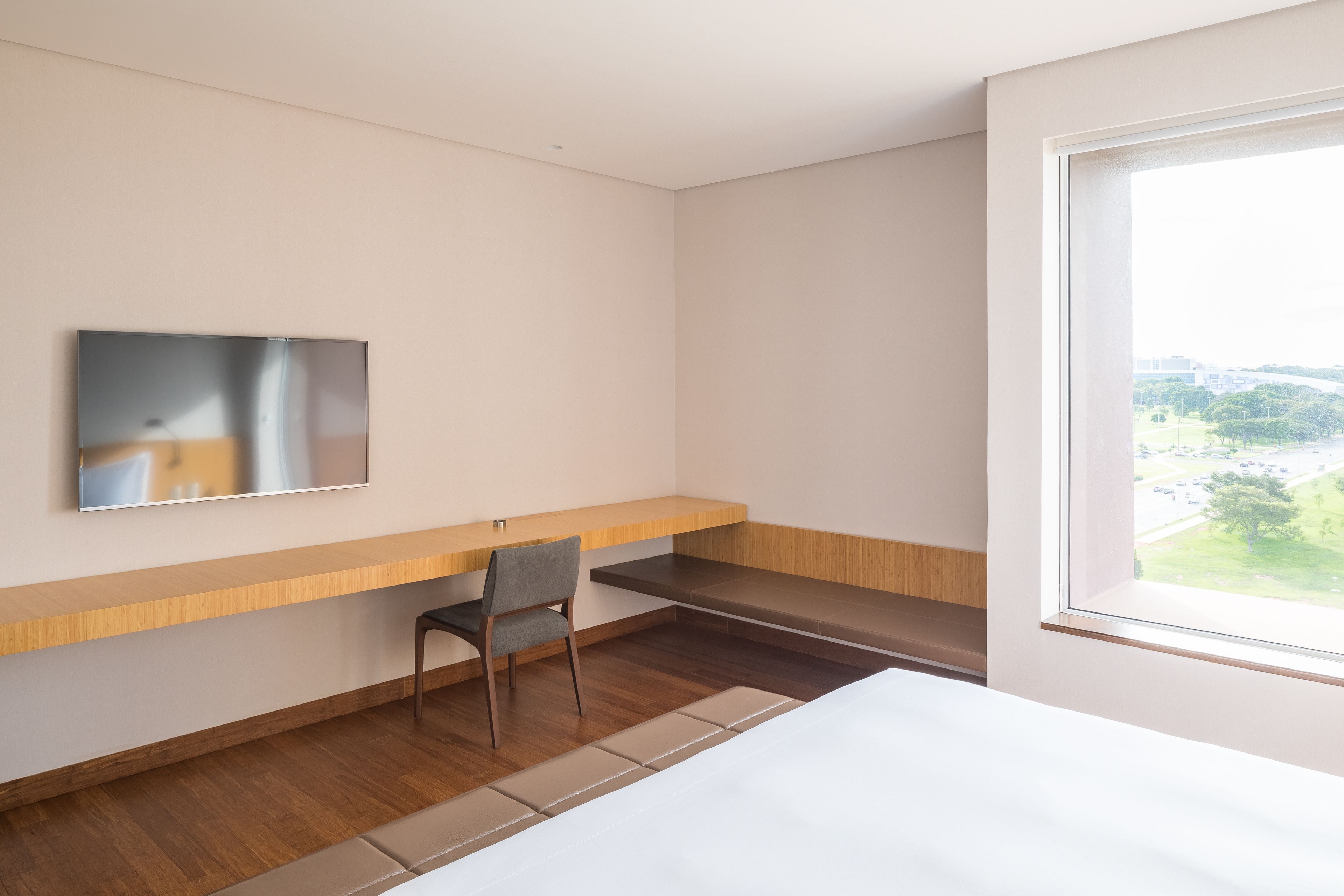 A modern hotel room with a bed, a desk, a chair, a TV mounted on the wall, and a window offering a view of the outdoors.