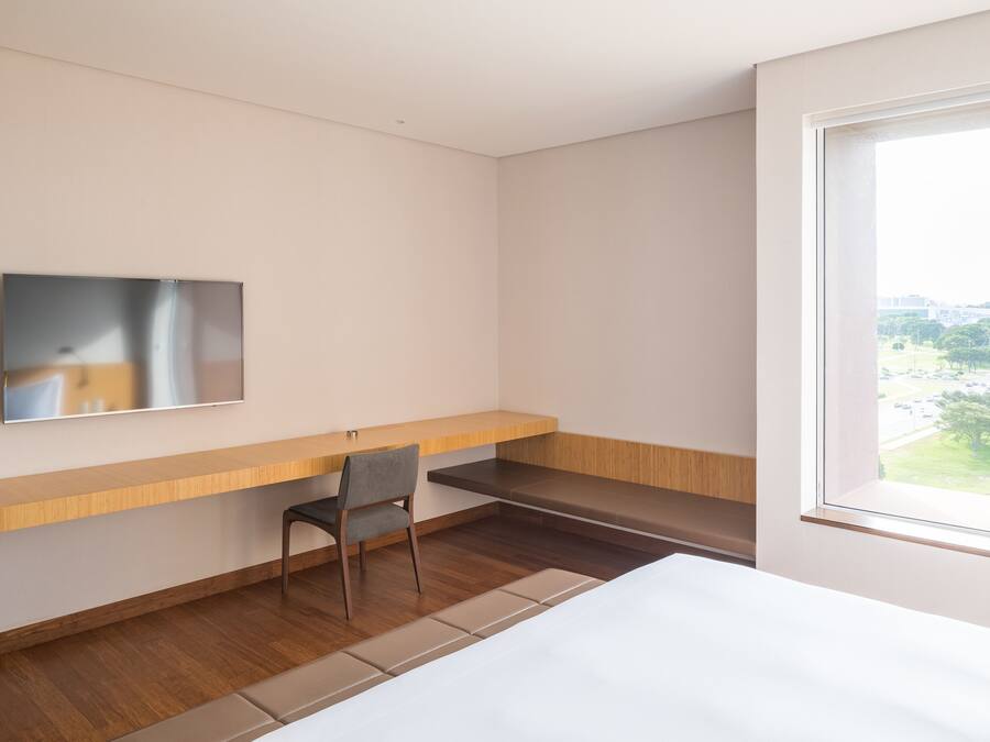 A modern hotel room with a bed, a desk, a chair, a TV mounted on the wall, and a window offering a view of the outdoors.