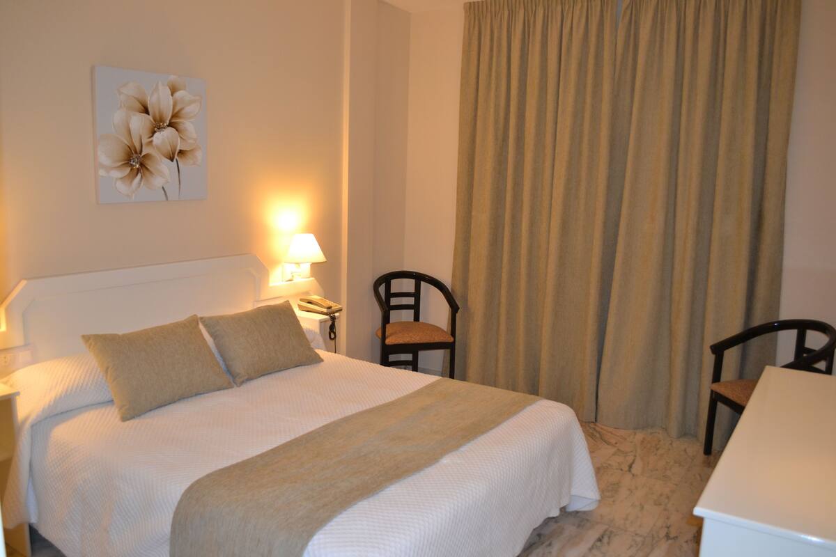 Habitación de hotel con una cama, dos sillas, un escritorio, una lámpara y un teléfono fijo en la pared.