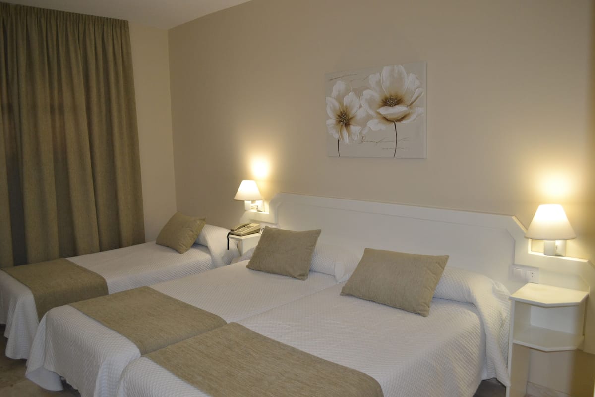 Habitación de hotel con dos camas, ropa de cama color beige, una obra de arte floral en la pared y lámparas de noche.