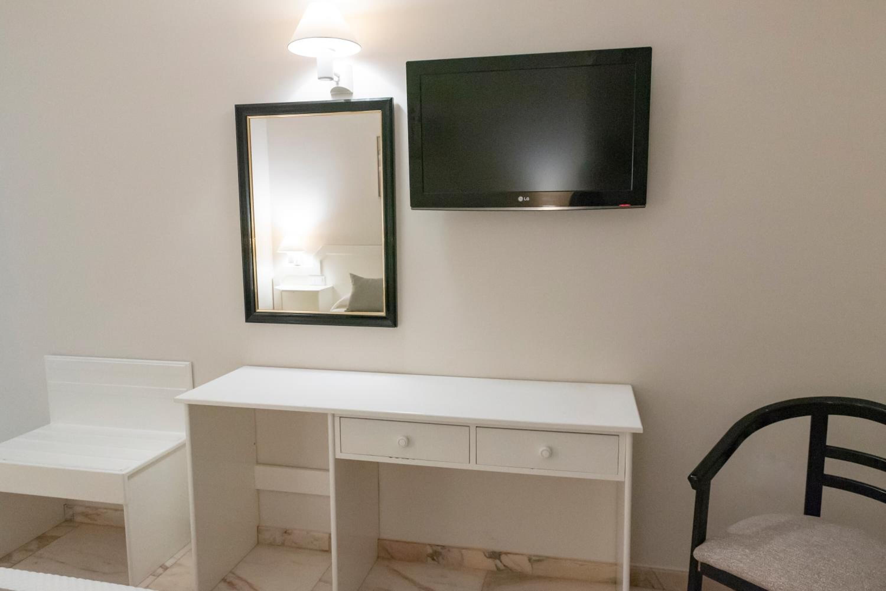 A white desk with drawers, a black chair, a framed mirror, and a flat-screen TV mounted on the wall.