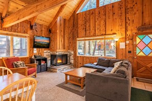 Interior acogedor de una cabaña de madera con chimenea, un televisor montado en la pared y un ventanal que ofrece vistas al exterior.
