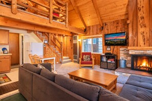 Interior de una cabaña de troncos con sala de estar, comedor, escalera, chimenea y televisor montado en la pared.