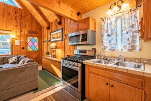 Un interior acogedor de cabaña con detalles de madera, una kitchenette con estufa y microondas, un sofá y una ventana con cortinas.