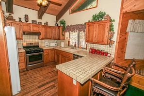 A kitchen with wooden cabinets, a tiled backsplash, a central island, and a dining area with a chair. - Summit Adventure (Big Bear Lake)
