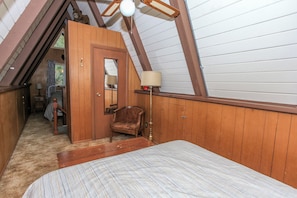 A bedroom with a sloped ceiling, wooden walls, a bed, a chair, and a lamp.