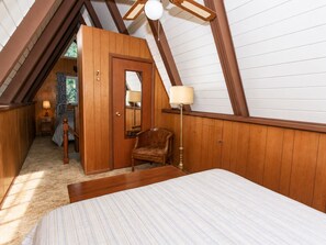 Una camera da letto con soffitto spiovente, un letto, una sedia, una lampada e una porta che dà su un'altra stanza.