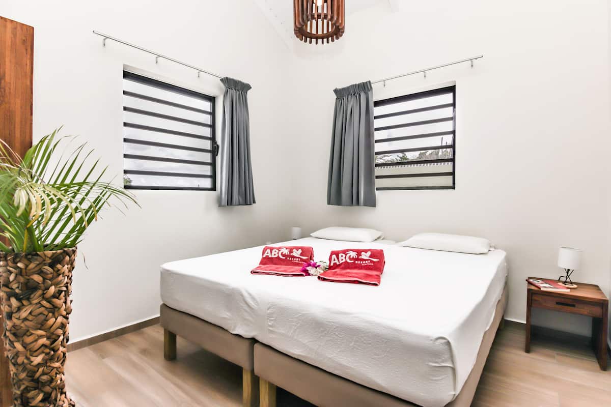 A neatly arranged hotel room with a bed, two windows with curtains, a bedside table with a lamp, and a potted plant.