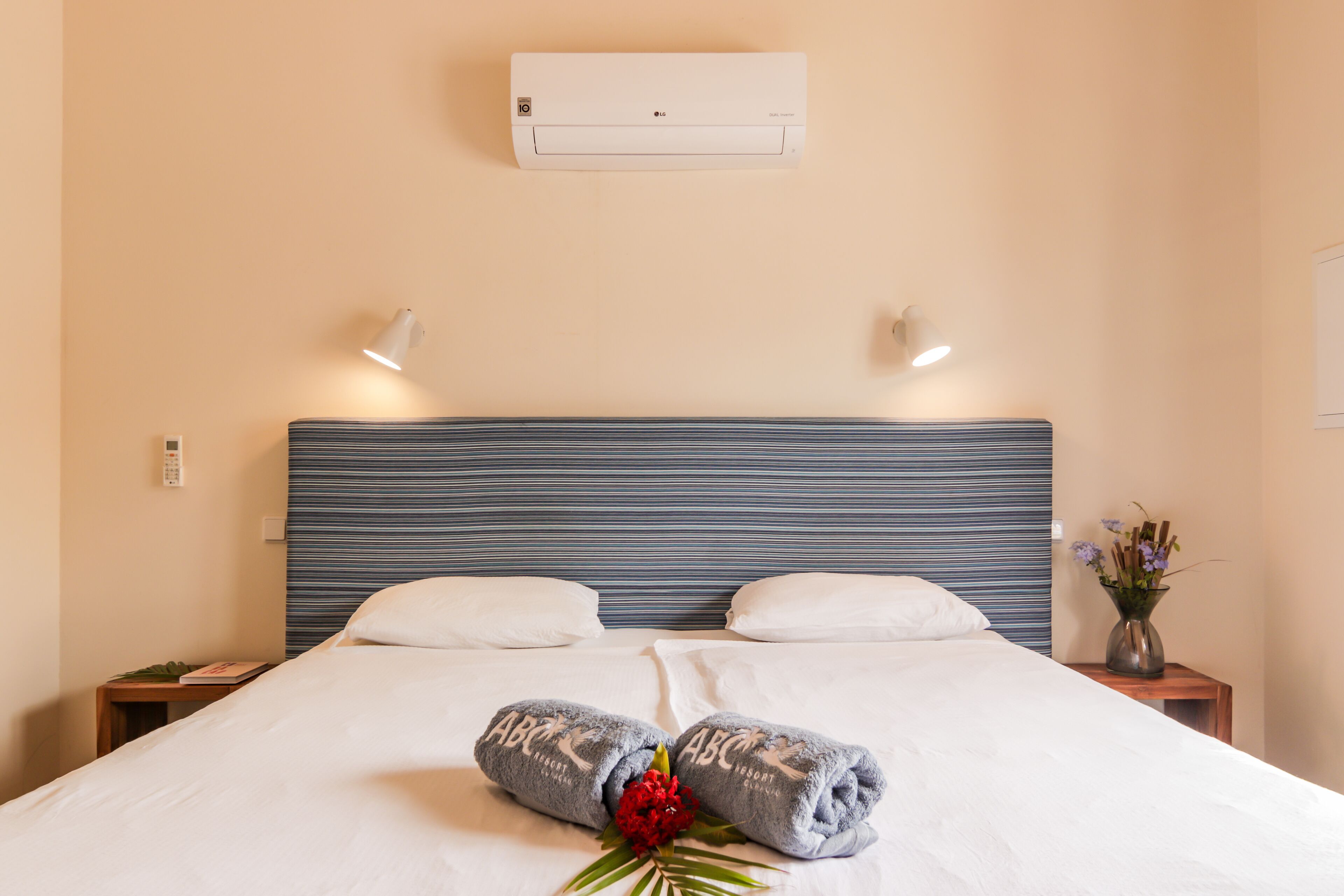 Een hotelkamer met een groot bed, twee kussens, een nachtkastje met een telefoon, een bijzettafeltje met een bloemenvaas en een airconditioning op de muur.