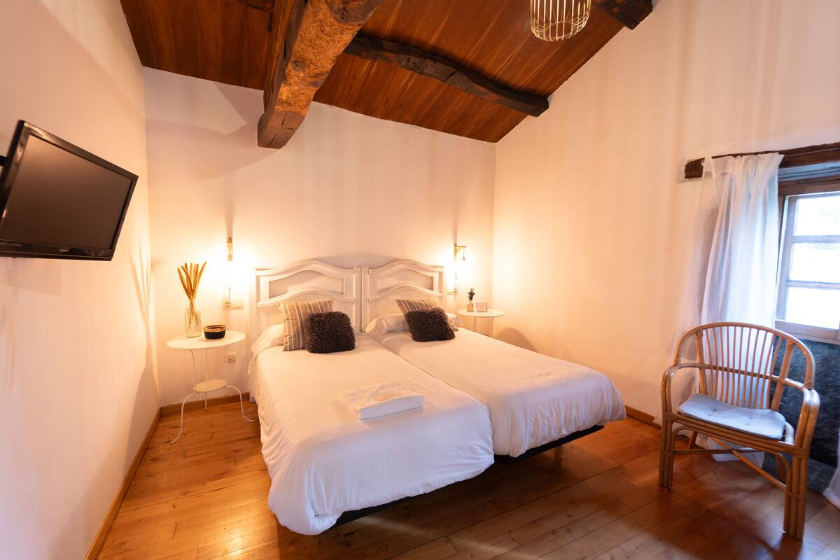 Ein Schlafzimmer mit zwei Betten, einem an der Wand befestigten Fernseher, einer Holzbalkendecke und einem Fenster mit Vorhängen.