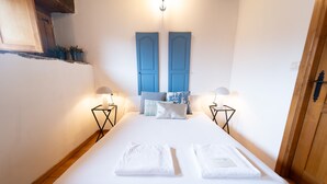 Un lit bien fait, avec du linge de lit blanc et deux serviettes, flanqué de deux lampes de chevet, dans une pièce au plancher et à la porte en bois.