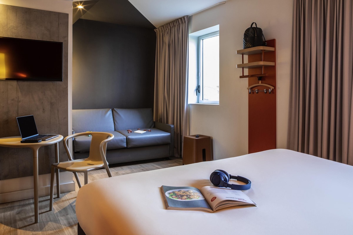 Ein modernes Hotelzimmer mit Bett, Schreibtisch mit Laptop, Stuhl, Sofa, Fenster mit Vorhängen und einem Wandregal mit einer Tasche.