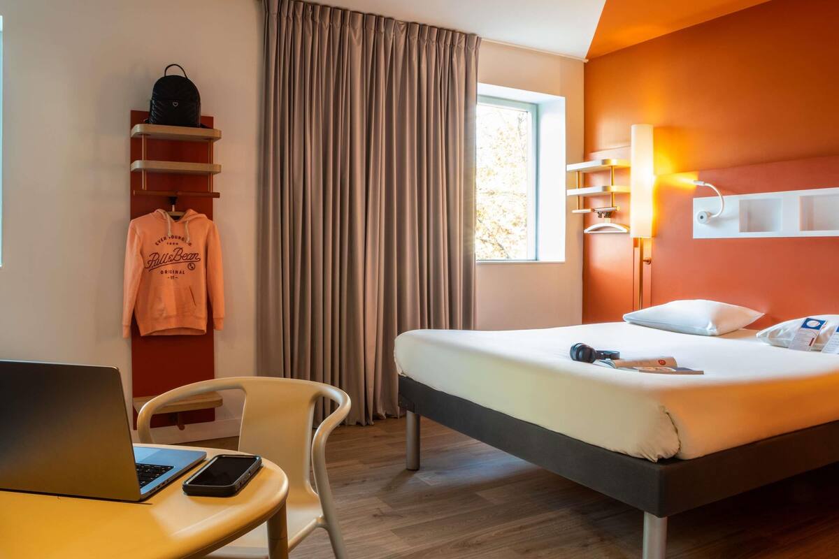 Habitación de hotel con una cama, un escritorio con una computadora portátil, una silla, un estante con una sudadera y una ventana con cortinas.