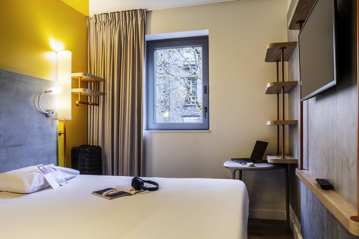 Habitación de hotel con cama, escritorio con una computadora portátil, ventana con vista a árboles y televisor montado en la pared.