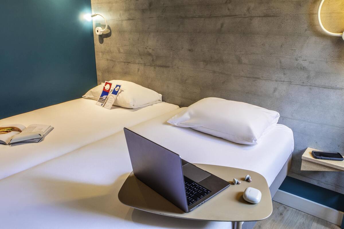 Una habitación de hotel moderna con dos camas, una laptop sobre una mesita y una mesita de noche con un teléfono y auriculares.