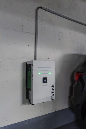 Eine weiße EVlink Ladestation mit grüner Kontrollleuchte, an einer Betonwand neben einem geparkten Auto angebracht.