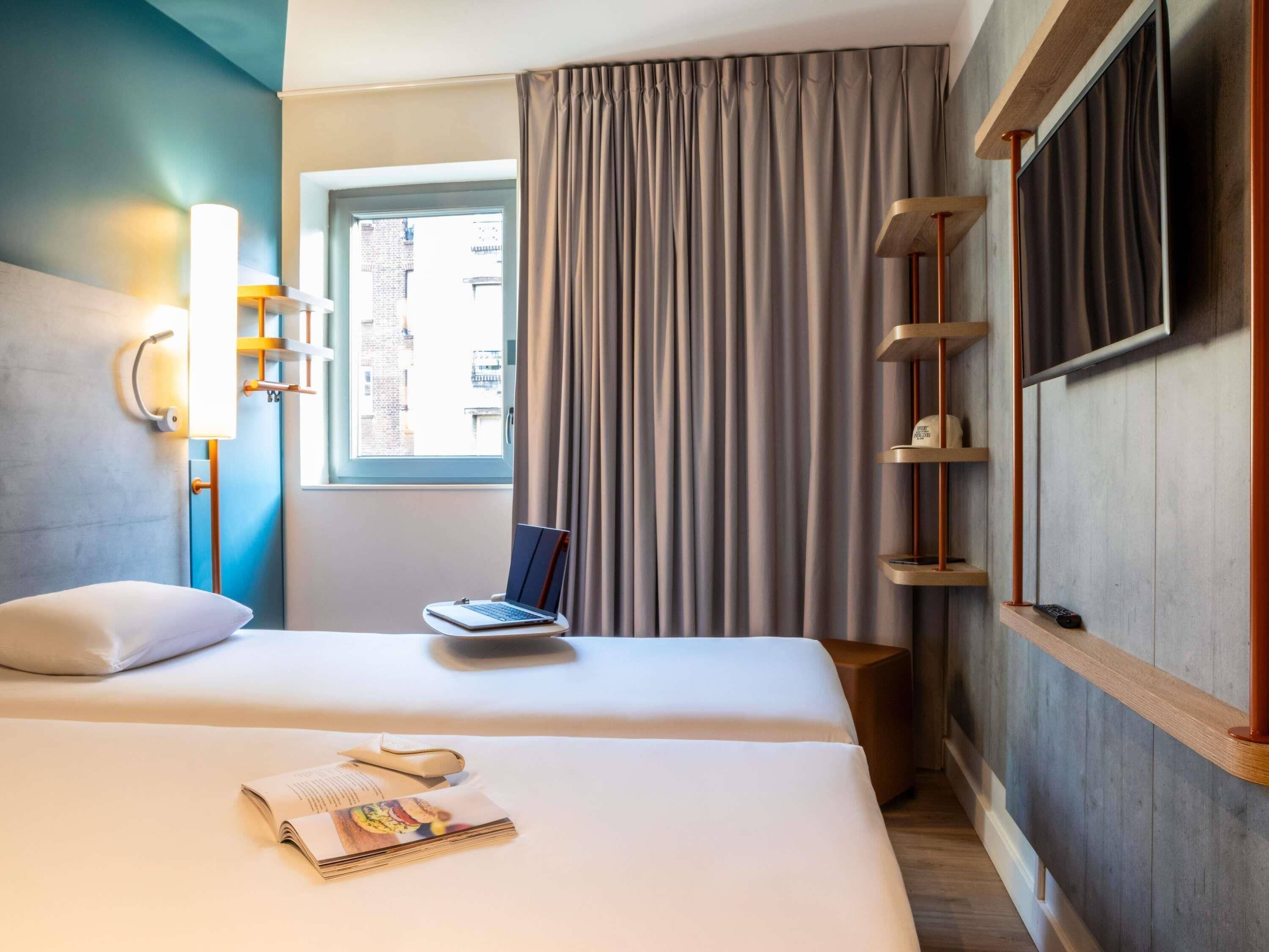 Habitación de hotel con cama, una laptop sobre la cama, ventana con cortinas, televisor montado en la pared y un estante con control remoto.
