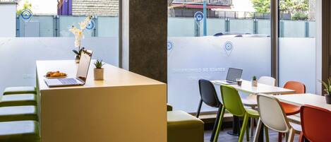 A modern office space with a white table, a laptop, and a potted plant. There are colorful chairs and a green bench.
