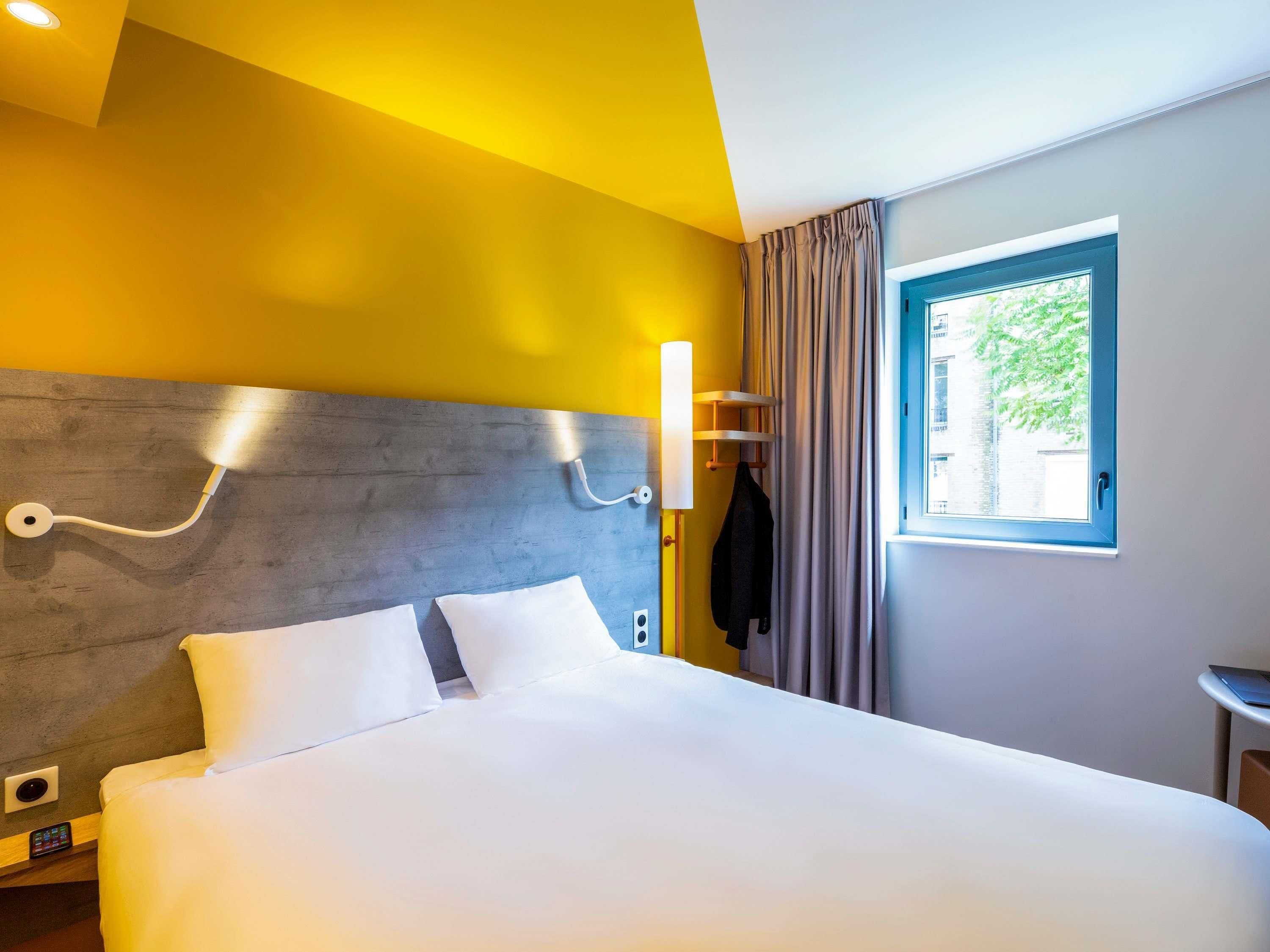 A hotel room with a bed, a headboard, a window with curtains, a wall-mounted light, and a coat rack.