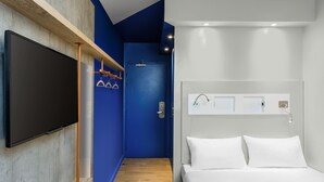 Ein modernes Hotelzimmer mit einem großen Flachbildfernseher, der an einer Holzhilfe angebracht ist, einer blauen Tür, einem weißen Bett mit zwei Kissen und einem kleinen Nachttisch mit einer Lampe.