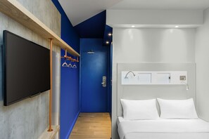 Une chambre d’hôtel moderne avec un grand téléviseur à écran plat fixé sur une étagère en bois, une porte bleue, un lit blanc avec deux oreillers et une petite table de chevet avec une lampe.