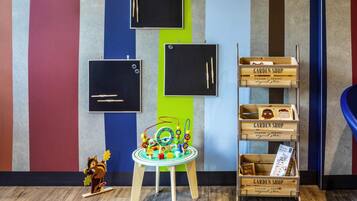 Una stanza con pavimento in legno, un tavolino con dei giocattoli, uno scaffale con cassette di legno etichettate 'GARDEN SHOP' e una parete con strisce verticali di diversi colori.