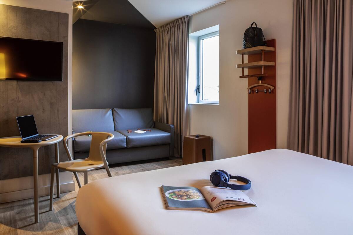 Una habitación de hotel moderna con una cama, un escritorio con una computadora portátil, una silla, un sofá, una ventana con cortinas y un estante montado en la pared con un bolso.
