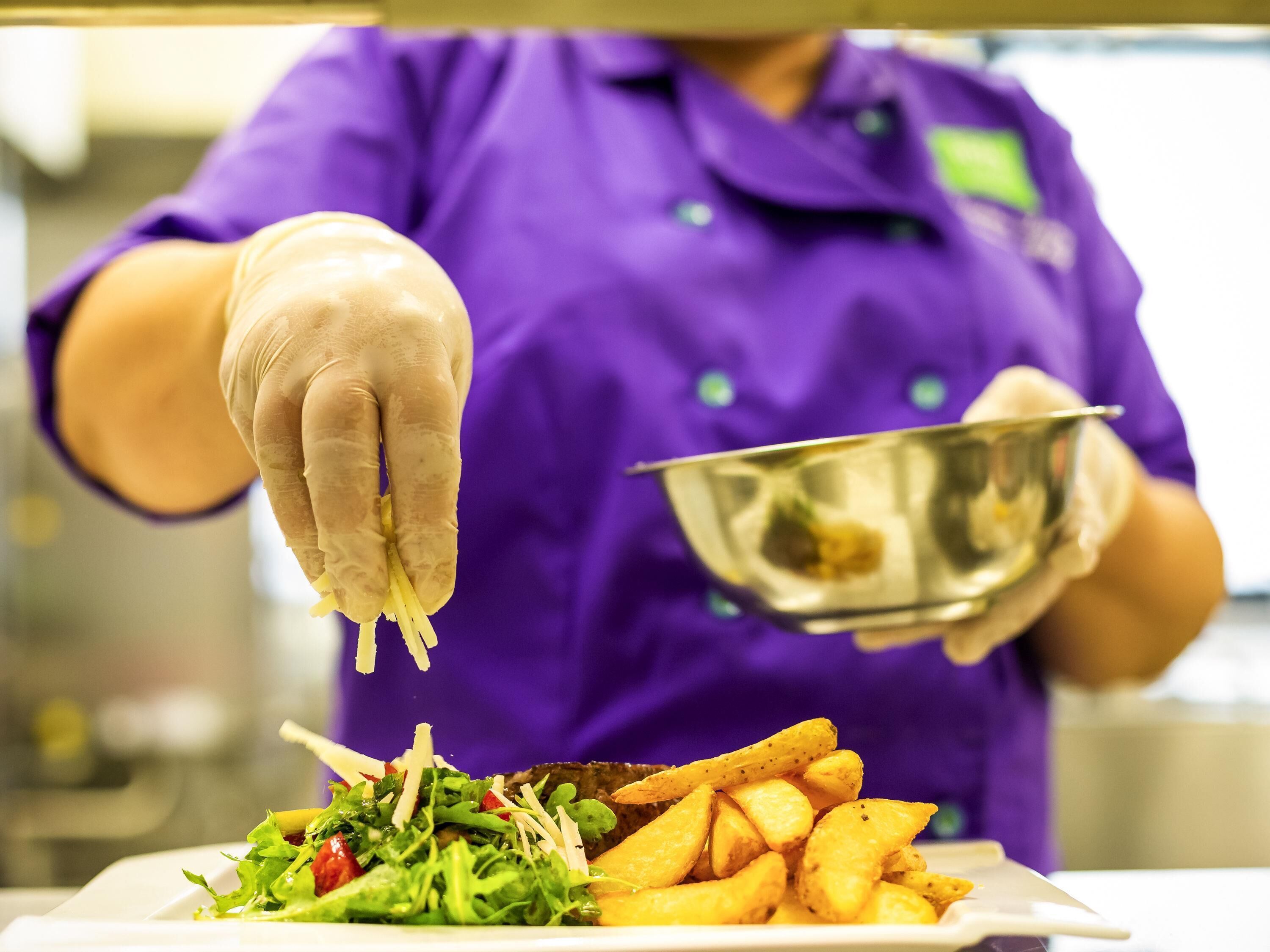A person in a purple uniform and gloves preparing food, with a plate of fries and salad in the foreground.
