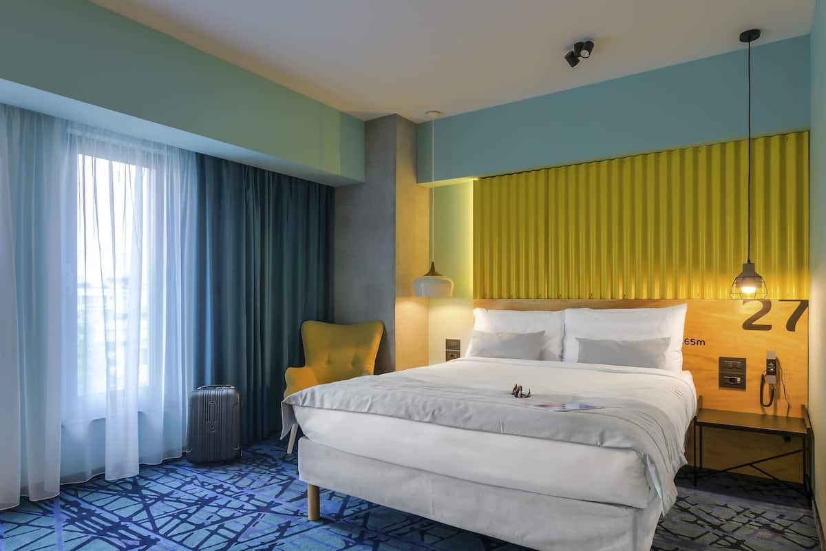 A hotel room with a bed, a chair, a lamp, a nightstand, and a window with curtains.
