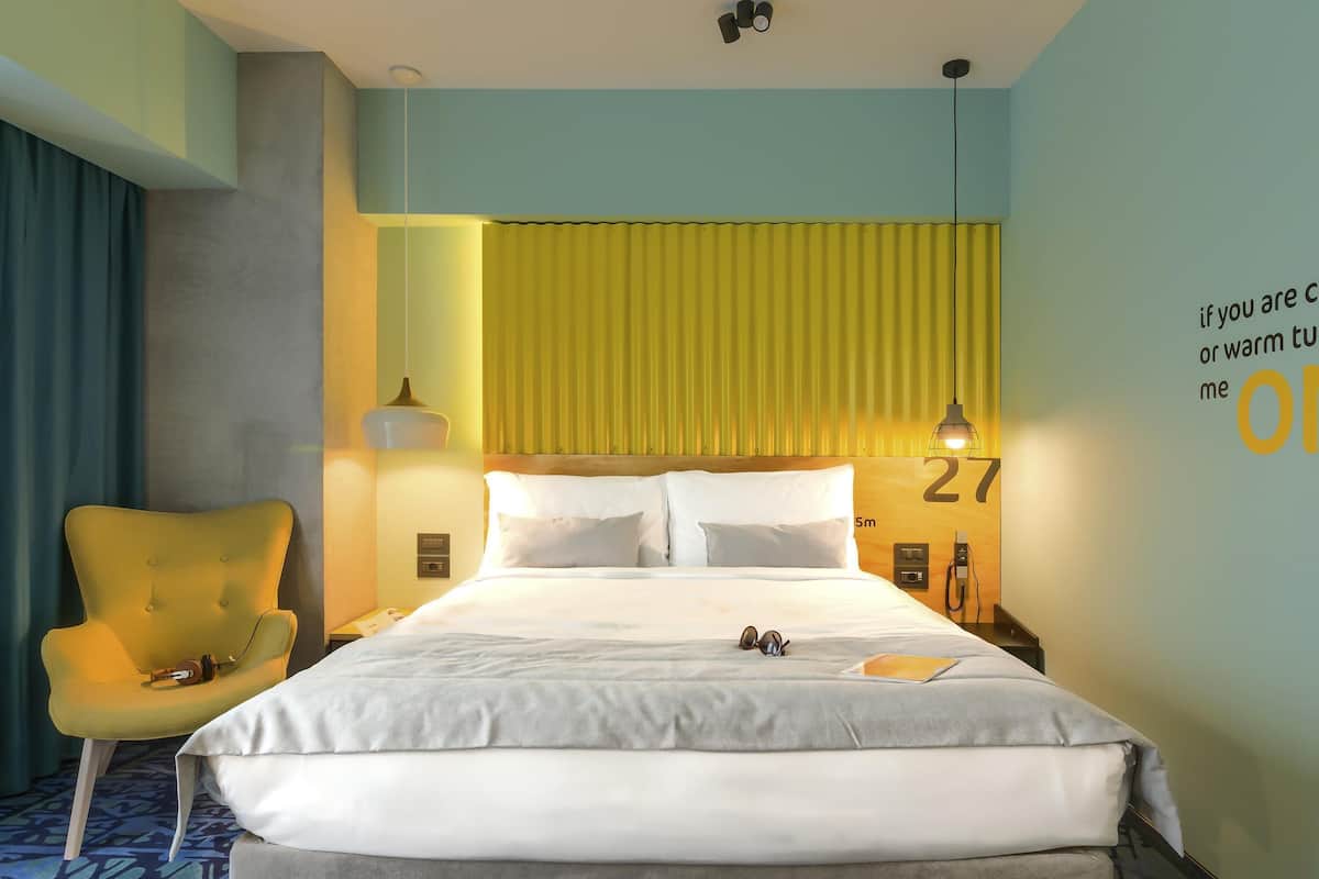 A hotel room with a bed, a chair, a wall clock, and a wall-mounted lamp.