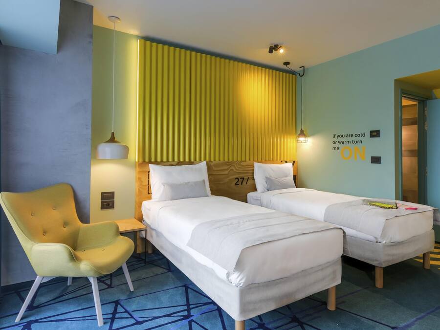 A hotel room with two beds, a chair, a desk, and a wall with a thermostat and a light switch.